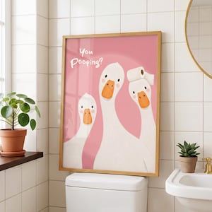 May include: A framed wall art print featuring three white geese against a solid pink background. The geese have orange beaks and one goose on the right wears a roll of white toilet paper on its head. The text "You Pooping?" is printed in white.