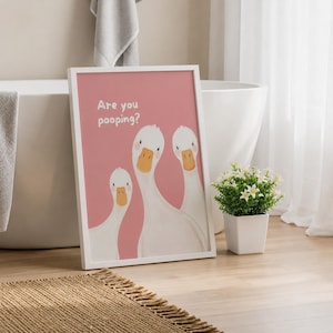 May include: A framed wall art print featuring a dusty pink background with three white cartoon ducks and the text Are you pooping? in white lettering. The ducks have orange beaks and small pink cheeks, arranged in a simple, clean composition.