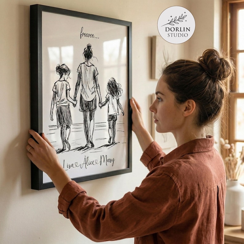 May include: A framed black and white sketch print depicting a mother and two children walking together, holding hands. The print includes the text "forever..." at the top and the names "Lisa & Alice & Mary" at the bottom. The print is being held by a person.