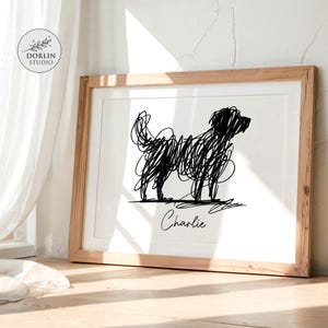 May include: A framed art print featuring a black scribble drawing of a dog on a white background. The dog's name, "Charlie," is written in cursive below the drawing. The frame is made of light-colored wood, and the print is from Dorlin Studio.
