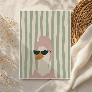 Pode incluir: Uma impressão de arte de parede retangular apresentando um pato branco com uma toalha rosa enrolada na cabeça e um roupão rosa correspondente. O pato tem um bico laranja brilhante e usa óculos de sol verde-escuros sobre listras verticais.