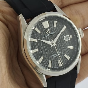 Op de afbeelding: Een roestvrijstalen Grand Seiko horloge met een zwarte textuur wijzerplaat en zilveren wijzers en markeringen. Het horloge heeft een datumvenster op 3 uur en een zwarte rubberen band.