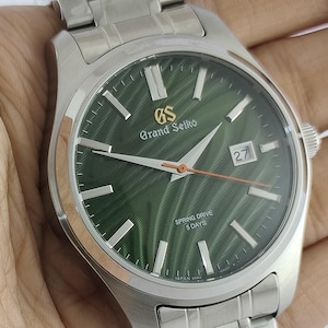 May include: A silver-tone stainless steel Grand Seiko watch with a textured green dial, silver hour markers, and a date window at the three o'clock position. The dial features the text GS Grand Seiko, Spring Drive, 5 Days, and Japan.