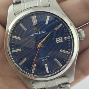 May include: A stainless steel Grand Seiko watch with a blue dial. The watch features a date display at the 3 o'clock position and the text "Spring Drive 5 Days". The watch has a silver strap and a silver case.