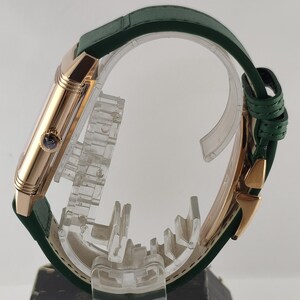 Puede incluir: Una vista lateral de un reloj de pulsera rectangular con caja de oro rosa pulido y correa de cuero verde oscuro. El reloj presenta una peque&ntilde;a corona circular en el lateral con un detalle azul y un cierre desplegable de oro rosa a juego.