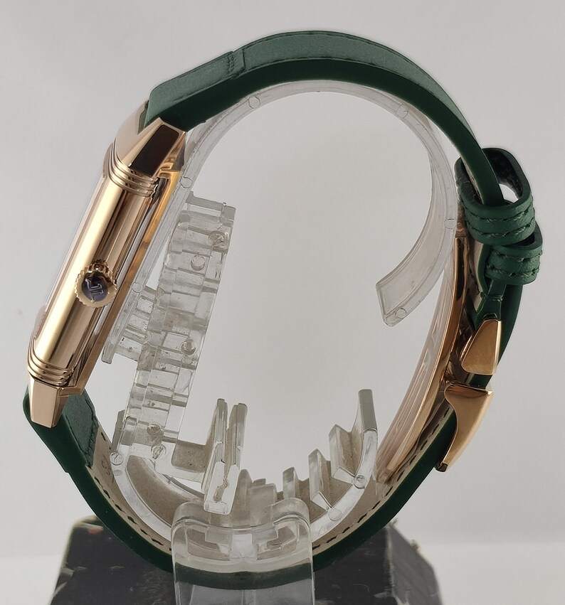 Puede incluir: Una vista lateral de un reloj de pulsera rectangular con caja de oro rosa pulido y correa de cuero verde oscuro. El reloj presenta una peque&ntilde;a corona circular en el lateral con un detalle azul y un cierre desplegable de oro rosa a juego.