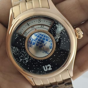 Puede incluir: Reloj de pulsera dorado con esfera negra con diseño de globo terráqueo y texto "U2". El reloj tiene una correa dorada y una esfera redonda con una visualización de la hora única.
