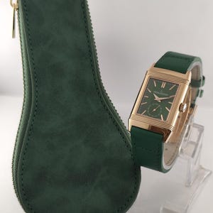 Puede incluir: Un reloj de pulsera rectangular en tono dorado con esfera verde y correa de cuero verde a juego, que presenta un subdial de segundos y marcadores dorados. Junto a &eacute;l, un estuche de almacenamiento verde oscuro en forma de gota con cremallera.
