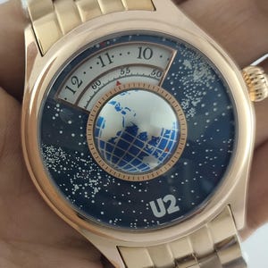 Puede incluir: Un reloj de pulsera dorado con un diseño único. La esfera del reloj presenta un fondo azul oscuro con puntos blancos, un globo terráqueo y el texto "U2". El reloj tiene una correa dorada y una esfera redonda.