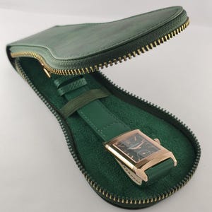 Puede incluir: Un reloj de pulsera rectangular con caja dorada y esfera negra, con correa de cuero verde, guardado en un estuche de viaje de cuero verde en forma de l&aacute;grima con cremallera de metal dorado y forro interior de color verde suave.