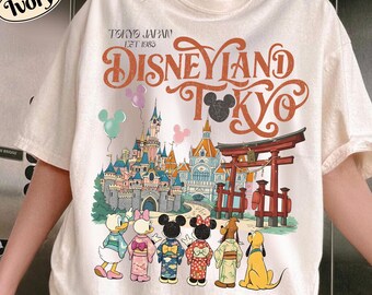 May include: An ivory t-shirt featuring a graphic print with the text "TOKYO JAPAN EST 1983 DISNEYLAND TOKYO". The design shows a castle, a red torii gate, and cartoon characters wearing traditional Japanese clothing, with balloons above them.