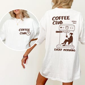 May include: White t-shirt with a brown "COFFEE CLUB" graphic and clock icon on the front. The back displays a brown illustration of a person making coffee, with the text "COFFEE CLUB SERVED DAILY EVERY MORNING."