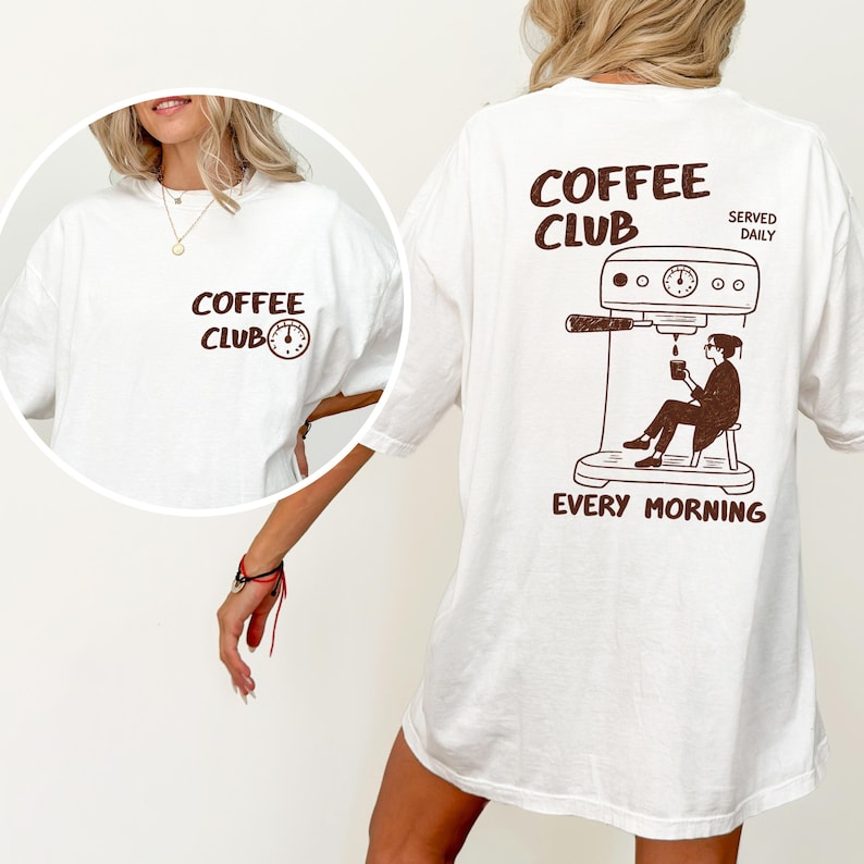 May include: White t-shirt with a brown "COFFEE CLUB" graphic and clock icon on the front. The back displays a brown illustration of a person making coffee, with the text "COFFEE CLUB SERVED DAILY EVERY MORNING."