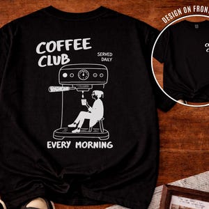 May include: Black t-shirt featuring a white illustration of a coffee machine and a person seated, holding a cup. The text "COFFEE CLUB" and "EVERY MORNING" are printed on the shirt. A smaller version of the design is on the front of the shirt.