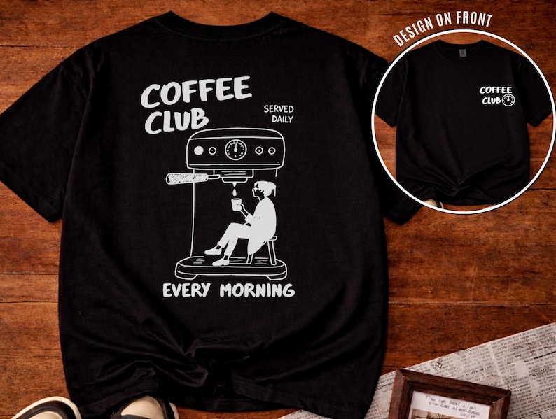 May include: Black t-shirt featuring a white illustration of a coffee machine and a person seated, holding a cup. The text "COFFEE CLUB" and "EVERY MORNING" are printed on the shirt. A smaller version of the design is on the front of the shirt.