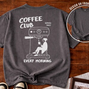 May include: Charcoal grey t-shirt featuring a white graphic of a coffee machine and a person enjoying coffee. The text "COFFEE CLUB SERVED DAILY EVERY MORNING" is printed on the shirt. A smaller image of the shirt is in the upper right corner.