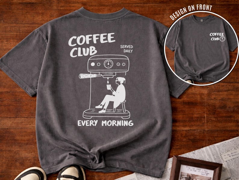 May include: Charcoal grey t-shirt featuring a white graphic of a coffee machine and a person enjoying coffee. The text "COFFEE CLUB SERVED DAILY EVERY MORNING" is printed on the shirt. A smaller image of the shirt is in the upper right corner.