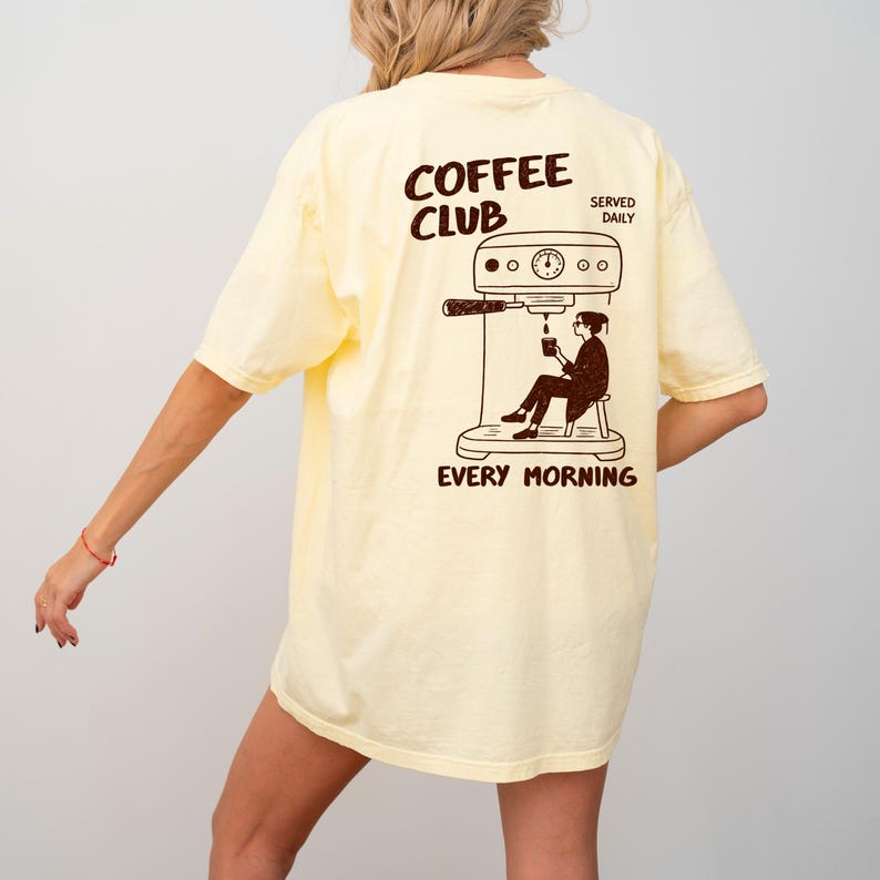 May include: A pale yellow t-shirt with a brown graphic on the back. The graphic shows the words "COFFEE CLUB" above a coffee machine illustration and a person drinking coffee. The words "EVERY MORNING" are below the illustration.