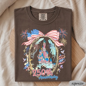 May include: A dark brown t-shirt featuring a graphic of a castle inside an oval frame, topped with a large American flag bow. The design includes fireworks, birds, and the text 250th Anniversary in blue and pink. The shirt has a Comfort Colors label.