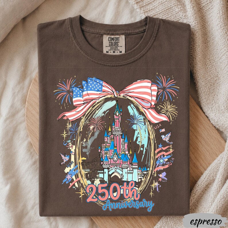 May include: A dark brown t-shirt featuring a graphic of a castle inside an oval frame, topped with a large American flag bow. The design includes fireworks, birds, and the text 250th Anniversary in blue and pink. The shirt has a Comfort Colors label.