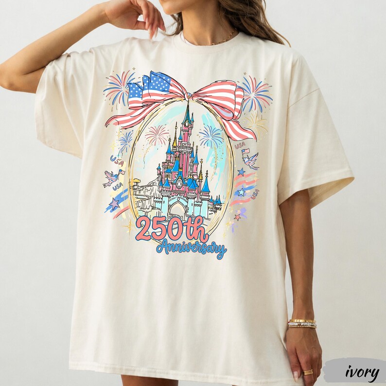 May include: An ivory t-shirt featuring a graphic of a castle inside an oval frame, topped with a large American flag bow. The design includes fireworks, the text 250th Anniversary, and small USA accents in red, white, and blue colours.