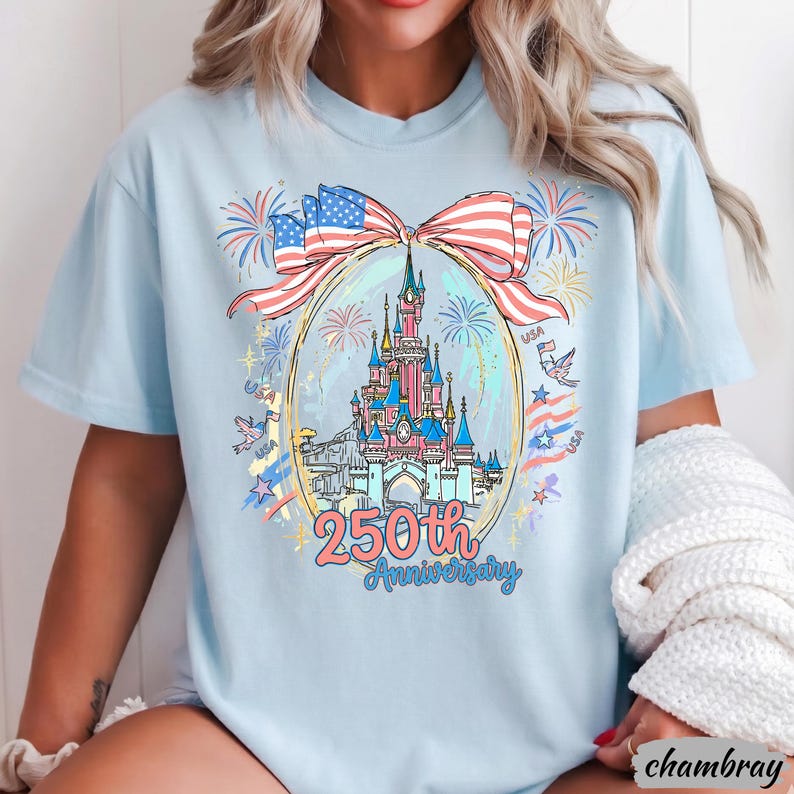 May include: A light blue t-shirt featuring a graphic print of a castle inside an oval frame, topped with a large American flag bow. The design includes fireworks, birds, and the text 250th Anniversary. The shirt has a crew neckline and short sleeves.
