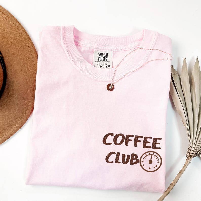 May include: A pale pink Comfort Colors t-shirt with the words "COFFEE CLUB" in brown lettering and a coffee gauge graphic. A rose gold necklace with a pendant is on the shirt. A brown hat and dried palm leaf are in the background.