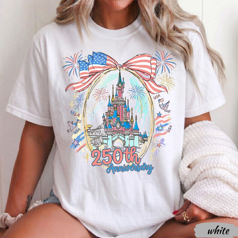 May include: A white t-shirt featuring a graphic of a castle inside an oval frame, topped with a large American flag bow. The design includes fireworks, birds, and the text 250th Anniversary in blue and pink, celebrating a patriotic theme.