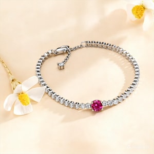 May include: A silver bracelet with a row of small, clear gemstones and a single, round, ruby-colored gemstone in the center. The bracelet is displayed on a light beige surface, with a white flower with a yellow center in the background.