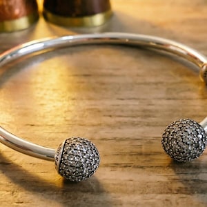 May include: A silver open-ended bangle bracelet featuring two spherical end caps encrusted with small clear stones. The bracelet includes two additional silver spacer beads, each engraved with the word Pandora, positioned along the smooth band.