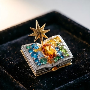 May include: A brooch shaped like an open book with a gold star rising from the center. The pages are encrusted with various round-cut gemstones in shades of blue, green, and yellow, featuring a large oval orange gemstone at the spine.