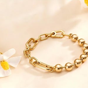 Könnte beinhalten: Ein goldfarbenes Armband mit einer Kombination aus Kettengliedern und kugelförmigen Perlen. Das Armband hat einen Verschluss und wird auf hellem Hintergrund mit einer Blume gezeigt.