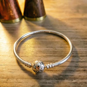 Puede incluir: Una pulsera rígida de plata pulida con un dije esférico central grabado con las letras P, A, N, D, O, R, A. La pulsera tiene una banda circular lisa con dos pequeñas cuentas de tope estriadas situadas a cada lado del dije central.