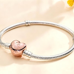 Può includere: Un bracciale d'argento con un ciondolo a forma di cuore in oro rosa. Il ciondolo presenta un testo inciso. Il bracciale ha un design a catena a serpente e una chiusura sicura. Gioielli.