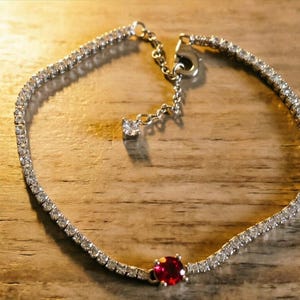 May include: A silver-toned tennis bracelet featuring a continuous row of small clear stones and a single prominent round red stone. The bracelet includes a thin chain extension with a small clear stone at the end and a lobster claw clasp.
