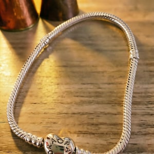 May include: A silver snake chain bracelet featuring a heart-shaped clasp. The clasp is engraved with the text PANDORA. The bracelet has a flexible, textured design with a polished finish, and the heart clasp serves as the primary closure mechanism.