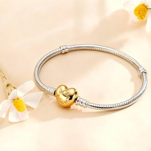 Puede incluir: Una pulsera plateada con un charm dorado en forma de corazón. El charm tiene la inscripción "Pandora". La pulsera está sobre un fondo beige claro con una flor blanca.