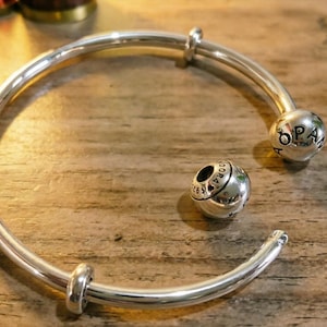 May include: A polished silver open bangle bracelet featuring a spherical end cap engraved with the text PANDORA and the number 925. A separate spherical charm with matching PANDORA 925 engraving sits inside the curve of the open metal bracelet.