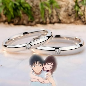 Puede incluir: Dos anillos de boda plateados con una pequeña gema transparente engastada en cada banda. Los anillos se muestran con una ilustración borrosa de estilo anime de una pareja abrazándose en el fondo. Los anillos son brillantes y reflectantes.