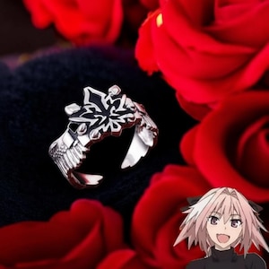 May include: A silver-colored ring with a black and silver floral design. The ring features wing-like details on the sides. The ring is set against a backdrop of red roses.