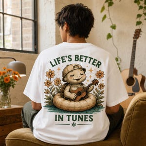 Puede incluir: Una camiseta blanca con un gráfico en la espalda de una tortuga de dibujos animados con gorra de béisbol, sentada en un flotador inflable mientras toca la guitarra. El texto "LIFE'S BETTER IN TUNES" está impreso en color verde oscuro.