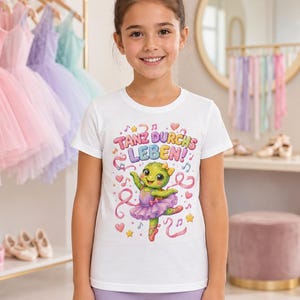 Op de afbeelding: Een wit T-shirt met korte mouwen en een kleurrijke afbeelding van een groene schildpad in een paarse tutu en balletschoenen. De tekst "TANZ DURCHS LEBEN!" staat erboven, omringd door muzieknoten, hartjes en sterretjes.