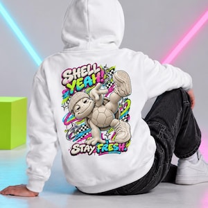 Könnte beinhalten: Ein weißer Kapuzenpullover mit einer Grafik auf der Rückseite, die eine Comic-Schildkröte mit Mütze und Turnschuhen zeigt. Das Design enthält den Schriftzug "SHELL YEAH!" und "STAY FRESH" in bunten Graffiti-Buchstaben, umgeben von Neon-Wirbeln und Sternen.