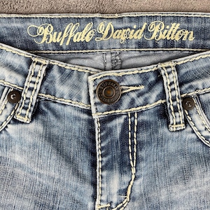 May include: Close-up of blue denim jeans with white stitching and a button closure. The brand name "Buffalo David Bitton" is embroidered in gold thread across the top. The jeans have a faded wash.