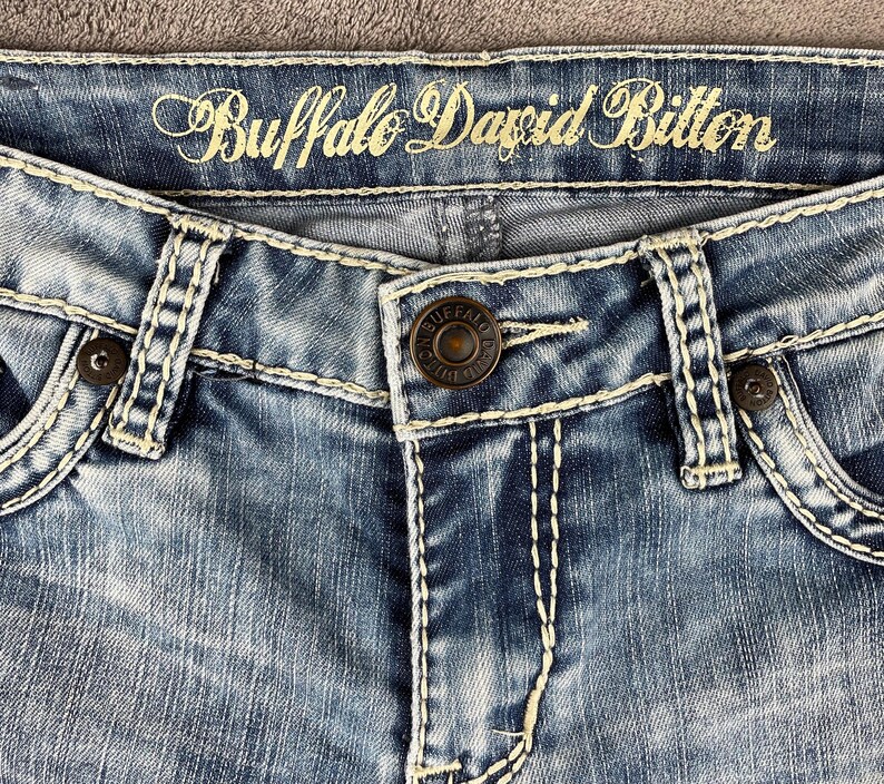 May include: Close-up of blue denim jeans with white stitching and a button closure. The brand name "Buffalo David Bitton" is embroidered in gold thread across the top. The jeans have a faded wash.
