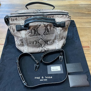 Może przedstawiać: Torebka ze wzorem skóry węża z czarnymi skórzanymi uchwytami i wykończeniami. Torba ma zapięcie na zamek błyskawiczny u góry i odpinany pasek na ramię. Torba leży na czarnej tkaninie z metkami "rag & bone NEW YORK".