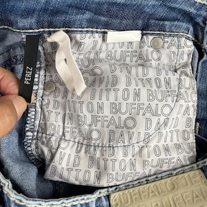 May include: Close-up of blue denim jeans with white stitching. The interior pocket lining features a repeating pattern of the words "DAVID BITTON BUFFALO". A black tag reads "PEREZ".