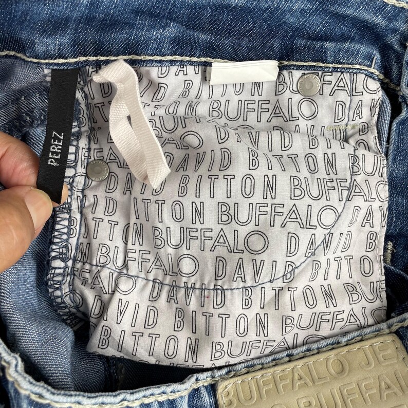 May include: Close-up of blue denim jeans with white stitching. The interior pocket lining features a repeating pattern of the words "DAVID BITTON BUFFALO". A black tag reads "PEREZ".