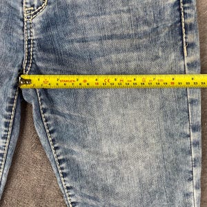 May include: Close-up of blue denim jeans with white stitching. A yellow tape measure shows a measurement of approximately 22 inches. The jeans are worn and faded, with visible wear marks.