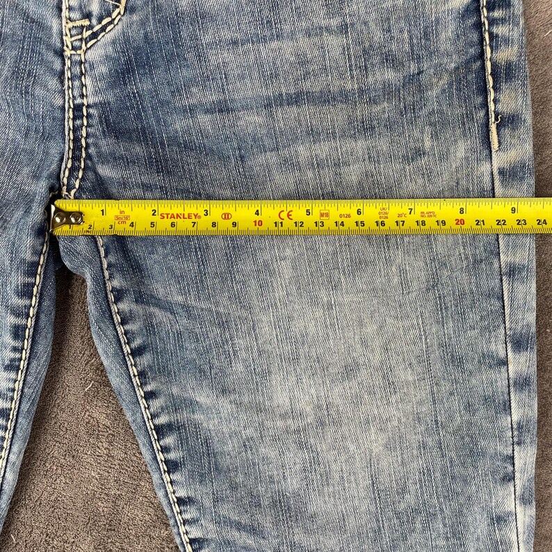 May include: Close-up of blue denim jeans with white stitching. A yellow tape measure shows a measurement of approximately 22 inches. The jeans are worn and faded, with visible wear marks.
